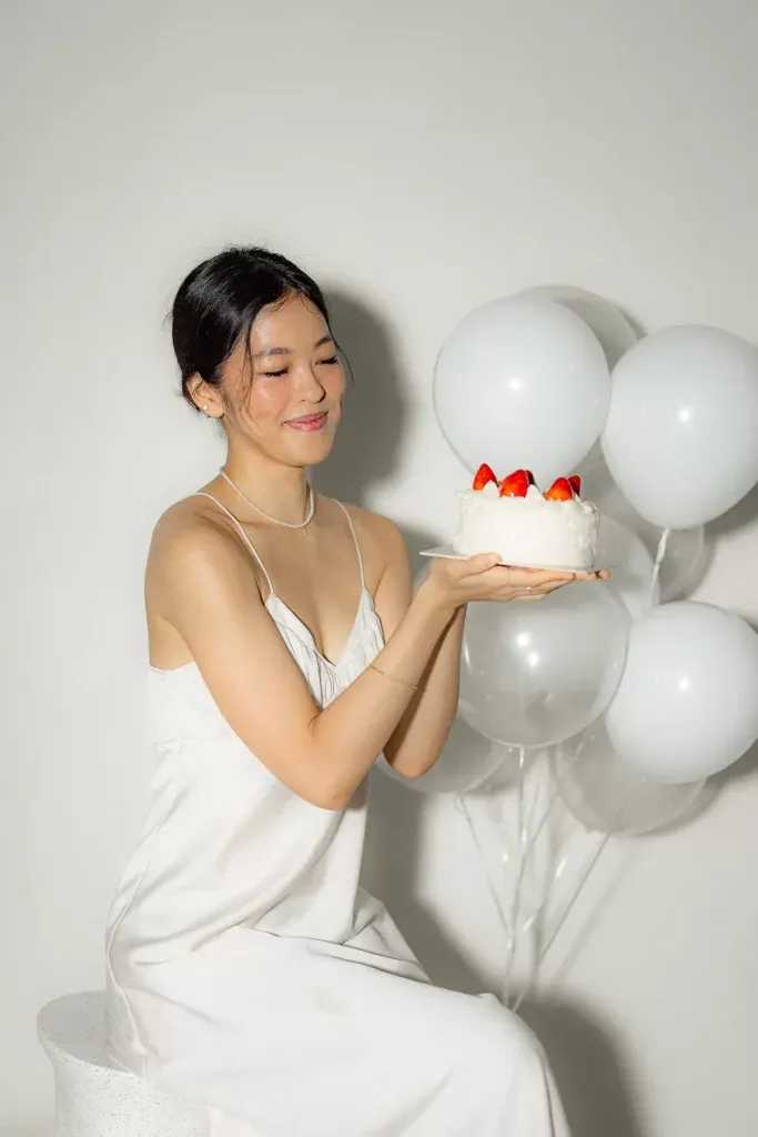 a girl in simple white dress holding a white cake and balloons