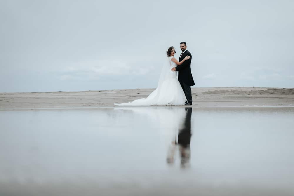 The Bride And Groom Walk Along The Beach Reflecting In water 