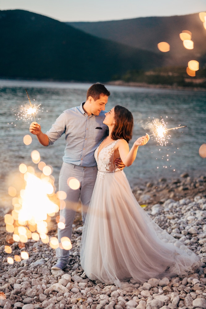 the bride and the groom hugging on the on the beach of the Mamula island and holding sparklers at the sunset