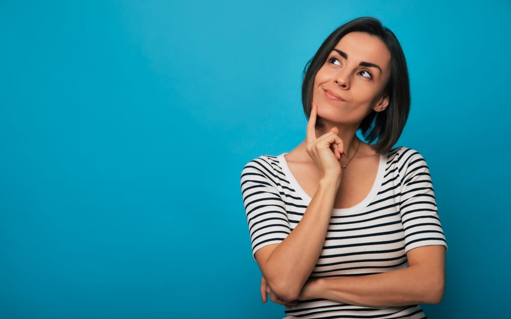 A young woman in a striped shirt stands against a blue background with a thoughtful expression, resting her finger on her chin.