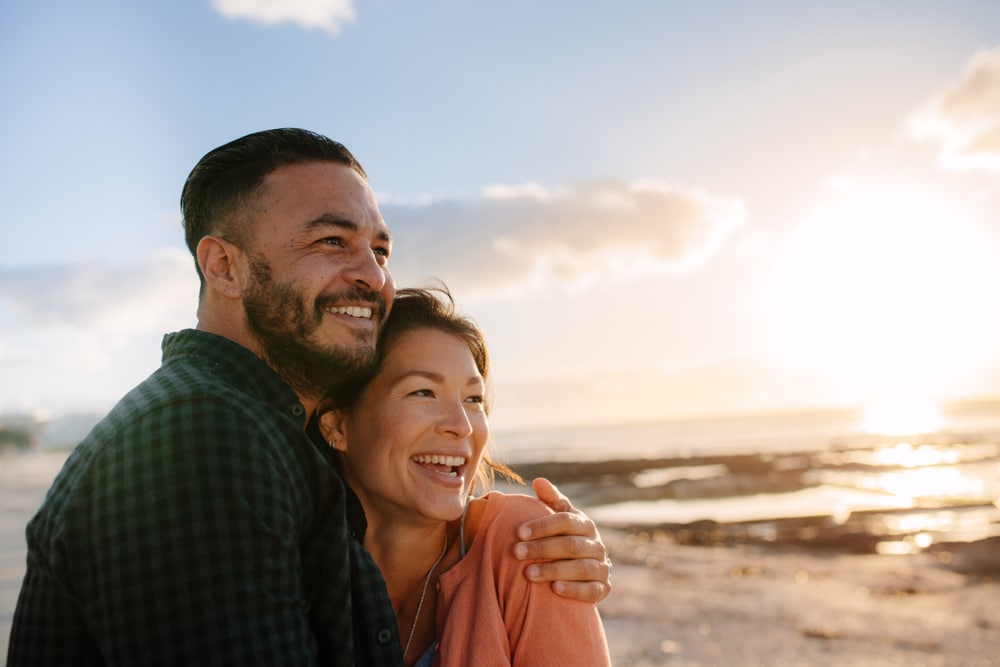 Man holding his partner with his arm around her standing at a beach and looking away.