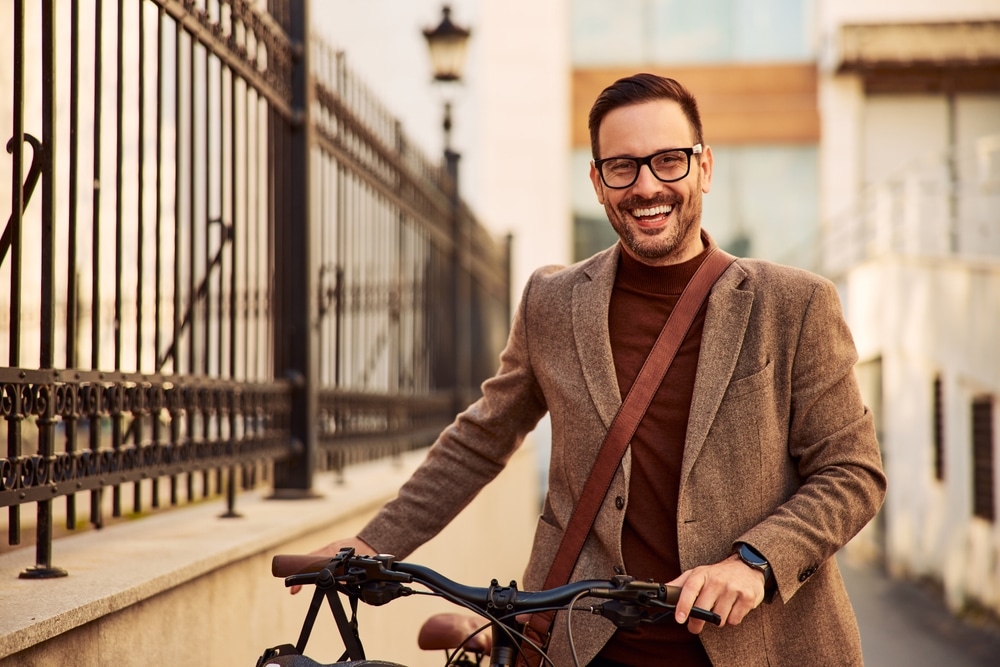 Portrait of a cheerful young adult high school teacher in a brown suit with glasses pushing his bicycle